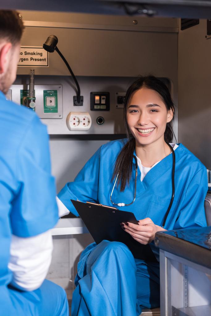 sonriente asiático paramédico sentado en ambulancia y mirando a la cámara
 - Foto, imagen