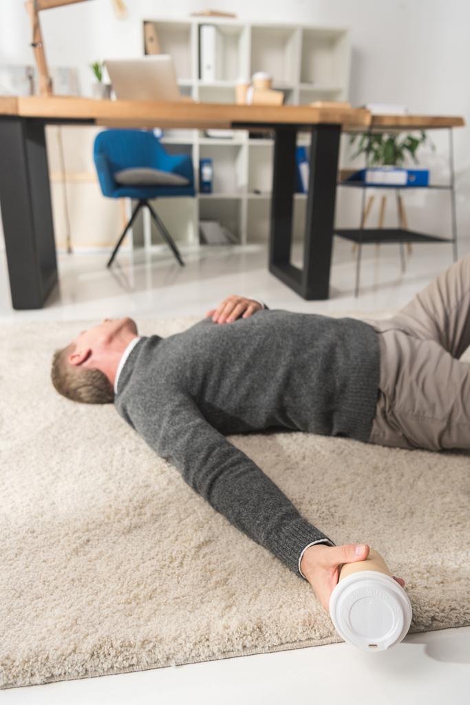 бессознательный мужчина лежит на полу в офисе и держит одноразовую чашку кофе в руке
 - Фото, изображение