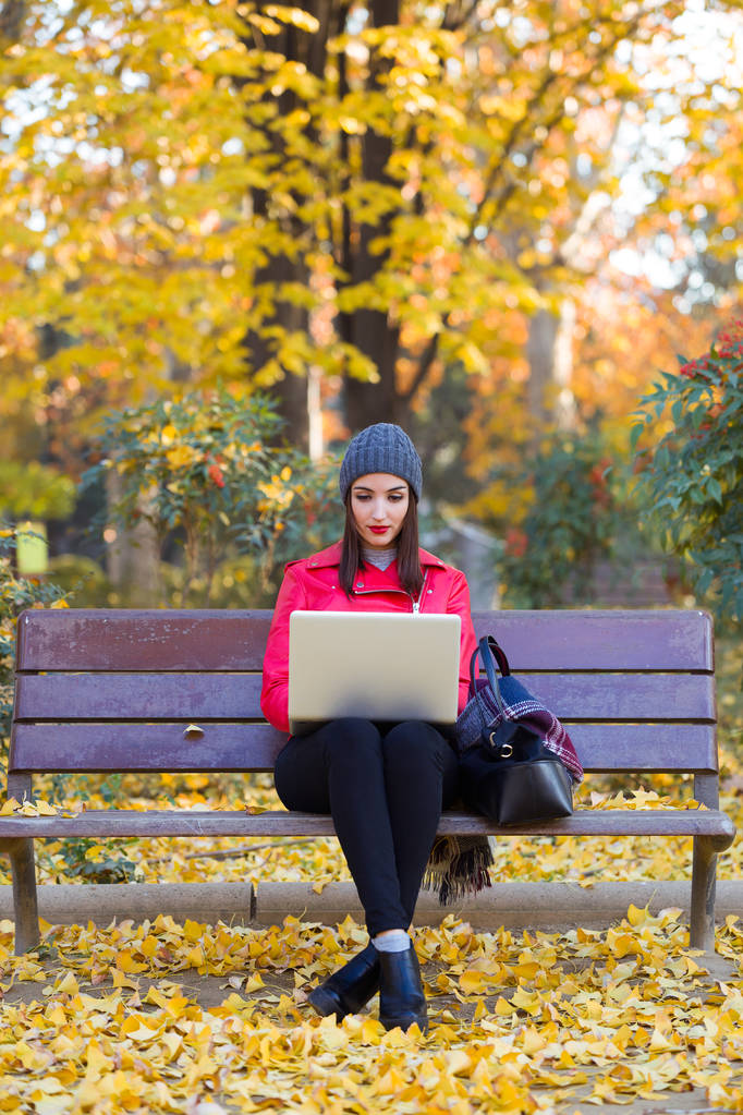 Hermosa joven sentada en un banco y usando su computadora portátil en otoño
. - Foto, imagen