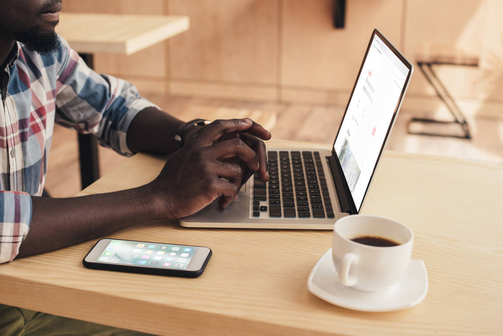 oříznutý pohled afroamerické muže pomocí přenosného počítače s web airbnb a smartphone v kavárně - Fotografie, Obrázek