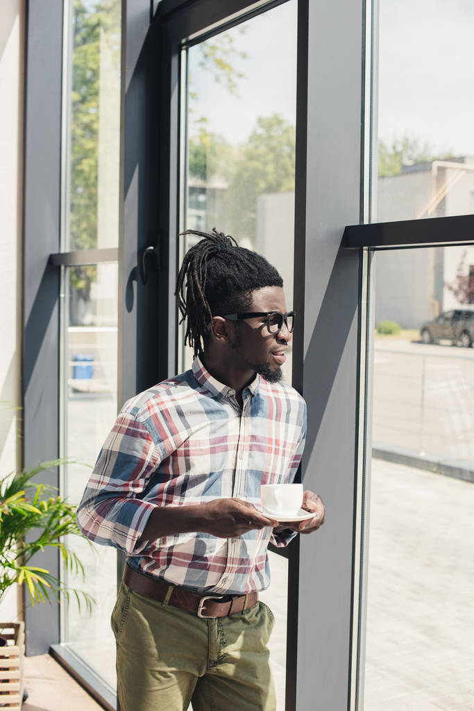 Αφρικανική αμερικανική άνθρωπος που πίνει καφέ ενώ στέκεται στο παράθυρο - Φωτογραφία, εικόνα