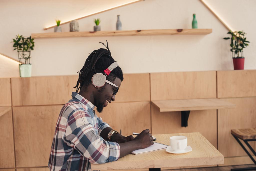 χαμογελώντας αφρικανική αμερικανική άνθρωπος ακούει μουσική και γράφοντας στο Σημειωματάριο (Notepad) σε καφενείο  - Φωτογραφία, εικόνα