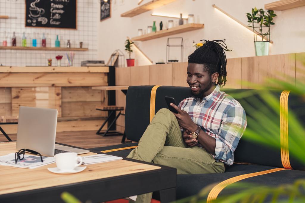 fröhlicher afrikanisch-amerikanischer Mann mit Smartphone und Laptop im Café - Foto, Bild