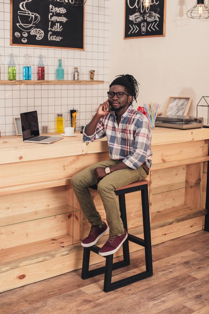 uomo afroamericano utilizzando il computer portatile e parlando su smartphone al bancone del bar nel caffè
 - Foto, immagini