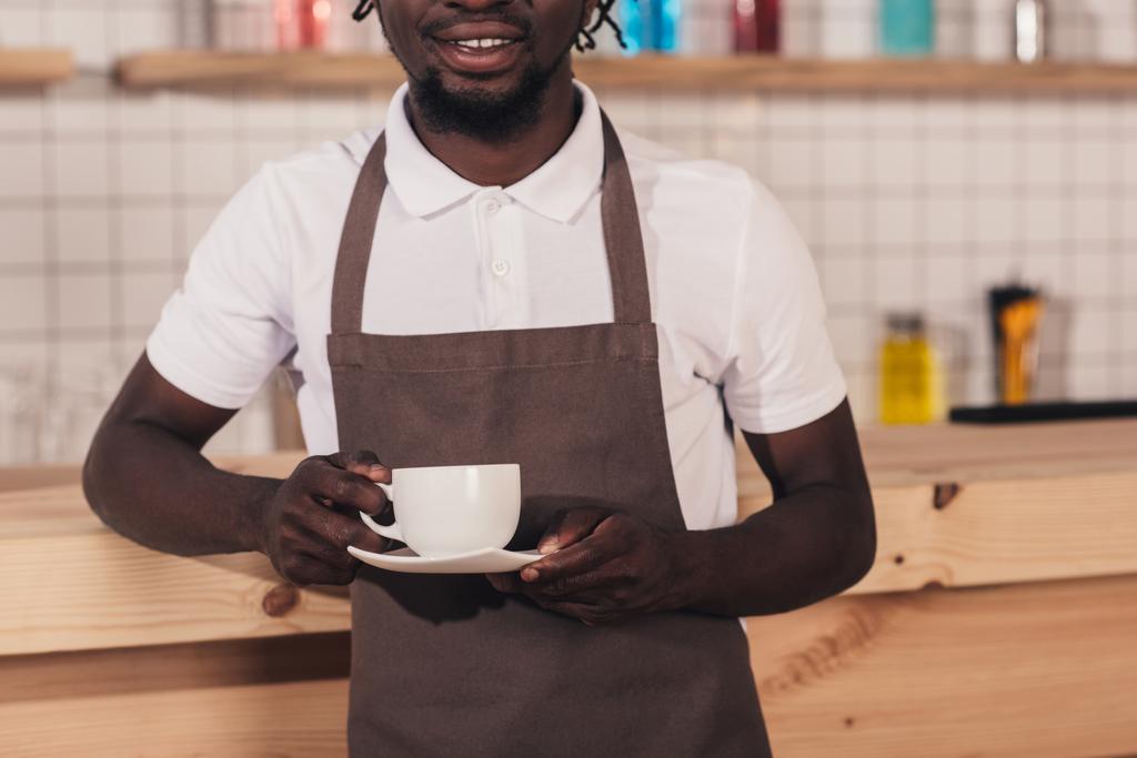bijgesneden weergave van Afro-Amerikaanse barista in schort houden van koffiekopje terwijl je op toog - Foto, afbeelding