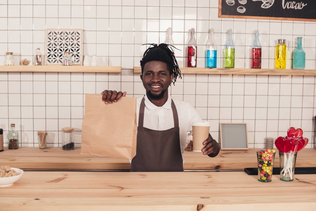 barista africano americano che tiene monouso tazza di caffè e pacchetto kraft al bancone del bar
 - Foto, immagini