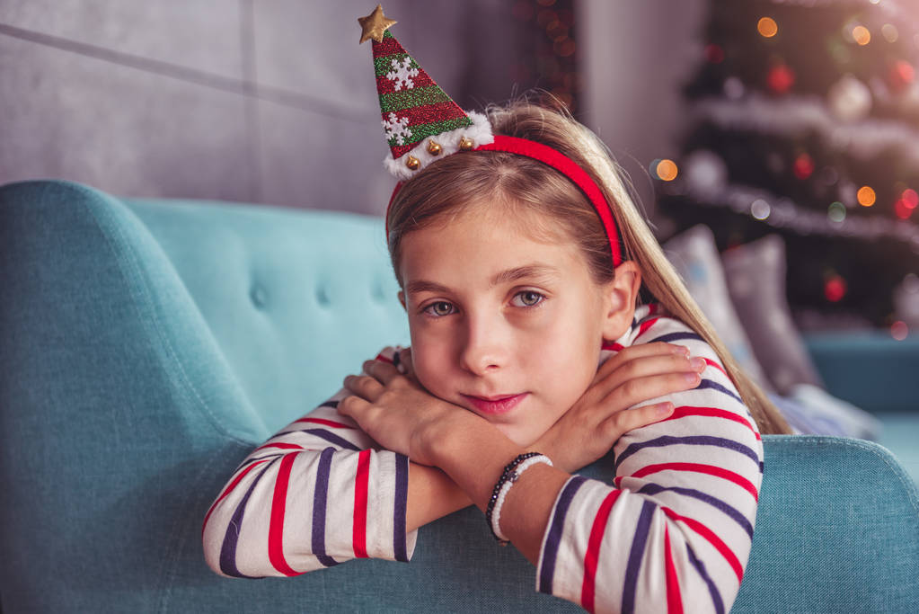 Маленькая девочка в шляпе Санты позирует на голубом диване на фоне рождественской елки
 - Фото, изображение