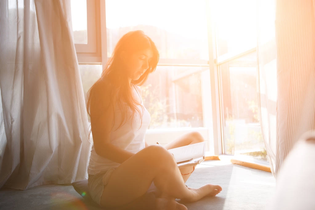 Φωτογραφία της μακρυμάλλης γυναίκα με βιβλίο κάθεται στο πάτωμα κοντά στο παράθυρο. - Φωτογραφία, εικόνα