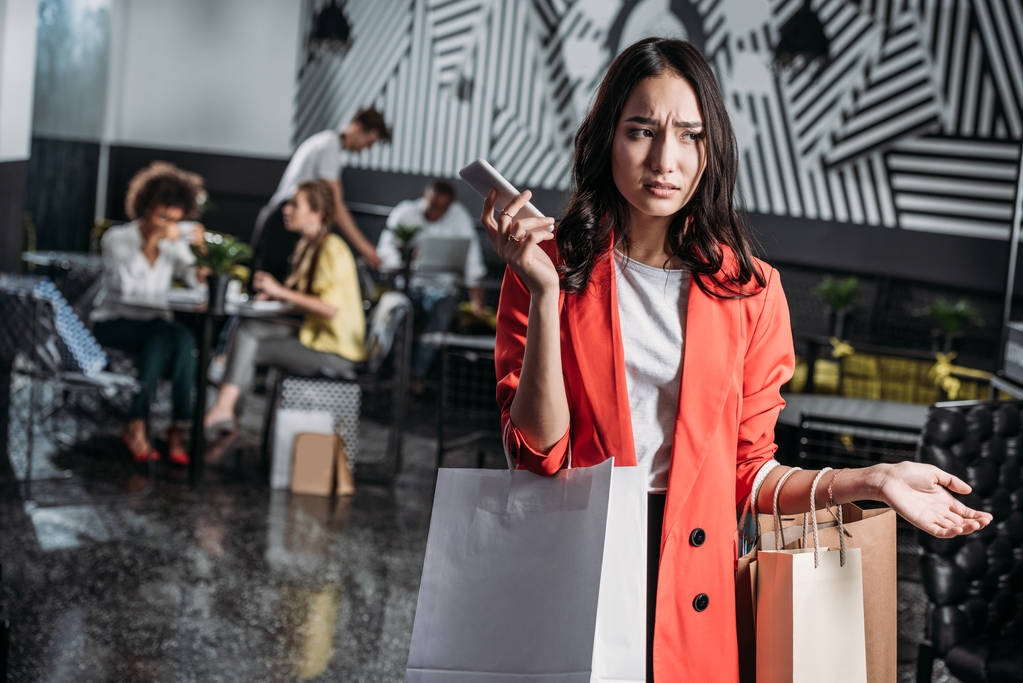 joven mujer frustrada con bolsas de compras y teléfono inteligente en la cafetería
 - Foto, imagen