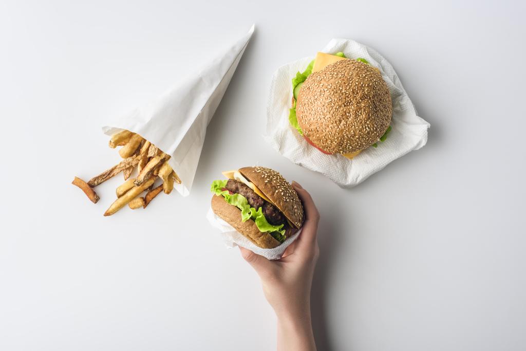 обрезанный вид женской руки с гамбургерами и картофелем фри в бумажном конусе, изолированный на белом
 - Фото, изображение