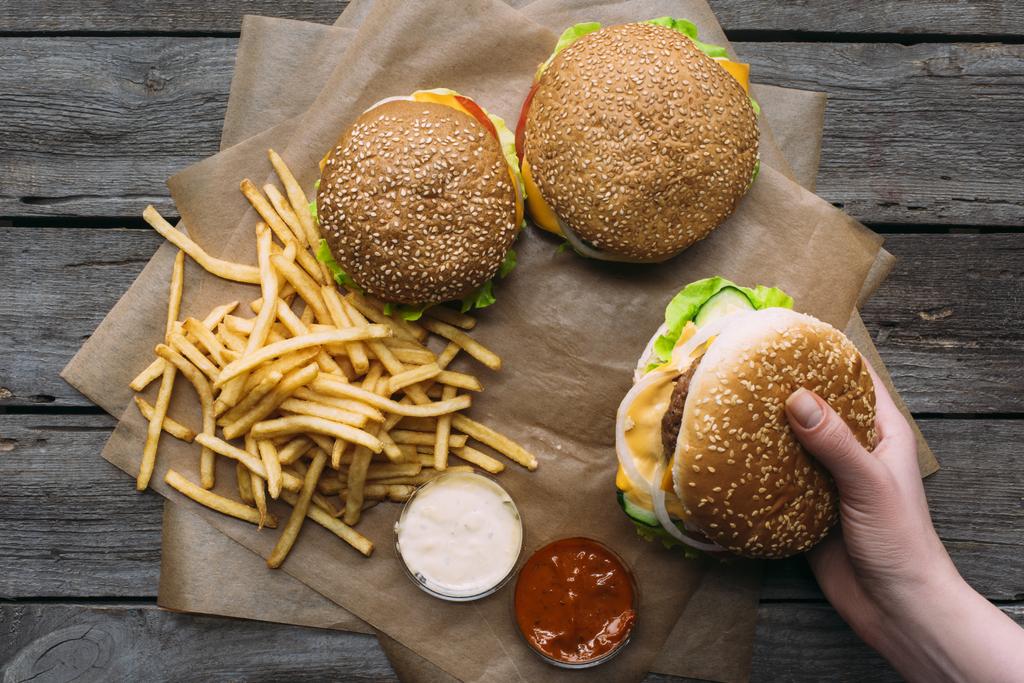 pohled shora ruky s hamburgery, hranolky a omáčky na pečicím papírem na dřevěnou desku - Fotografie, Obrázek