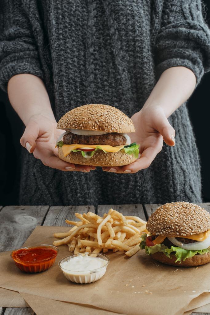oříznutý pohled ženy držící cheeseburger a držet u stolu s hranolky a omáčky - Fotografie, Obrázek