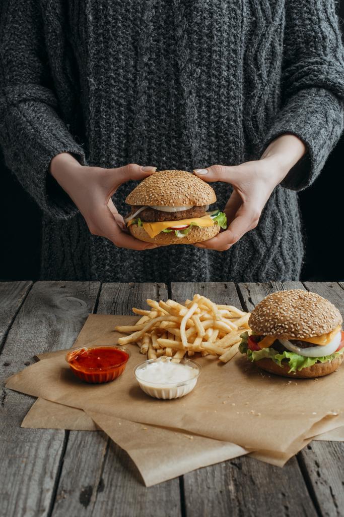 περικοπεί προβολή της γυναίκας κρατώντας cheeseburger και standig στο τραπέζι με τηγανητές πατάτες και σάλτσες - Φωτογραφία, εικόνα
