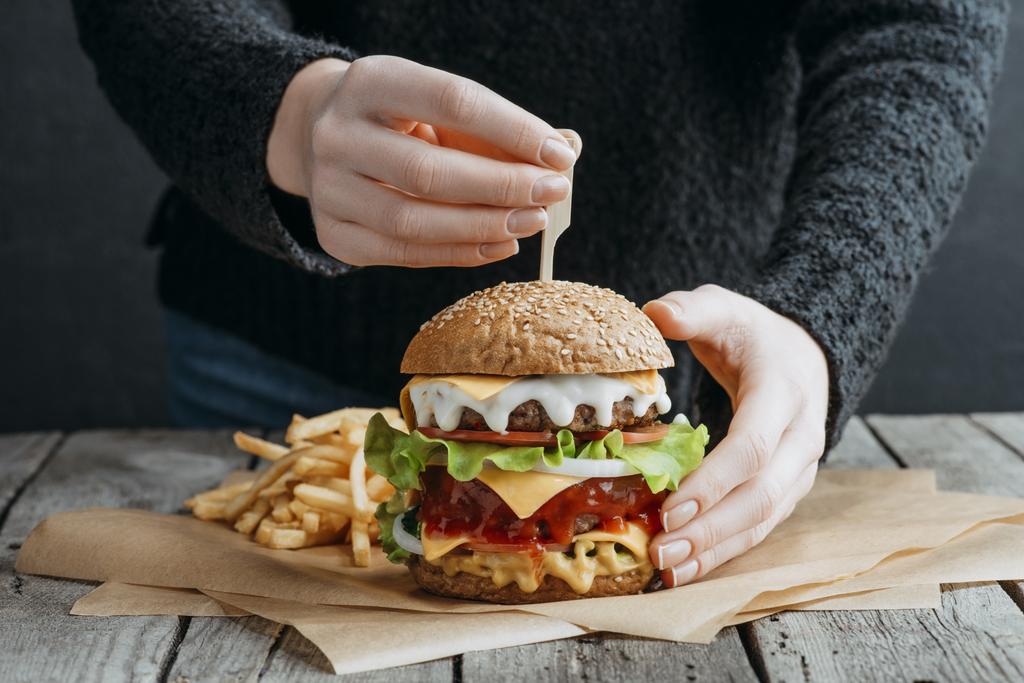 cropped view of female hands with big tasty cheeseburger and french fries on baking paper - Photo, Image