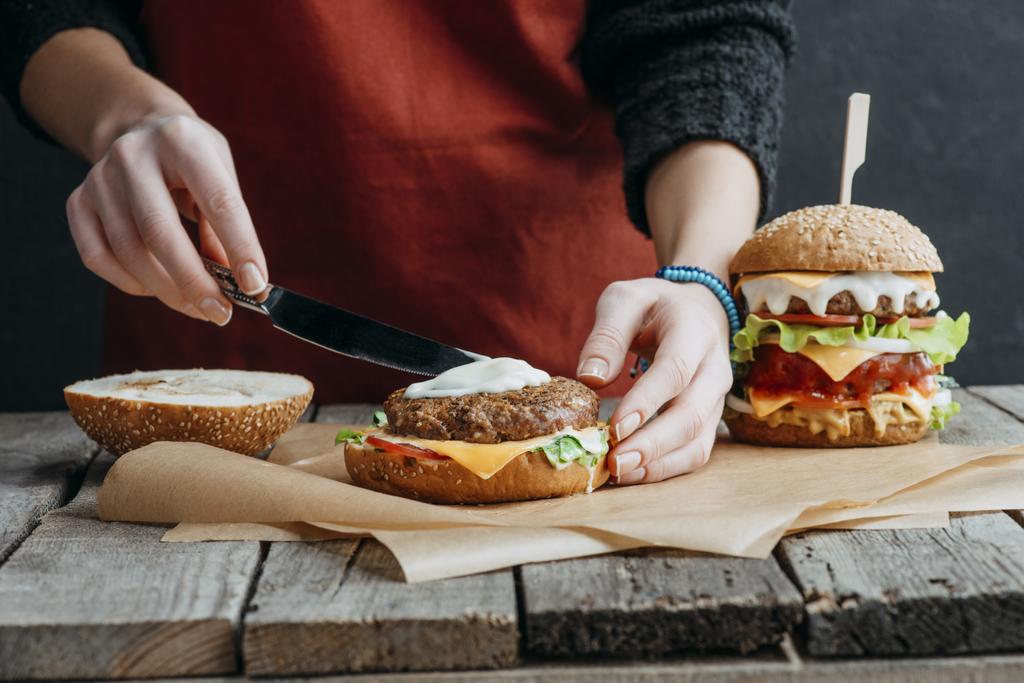 vista cortada da menina no avental fazendo saborosos cheeseburgers caseiros no papel manteiga na mesa de madeira
 - Foto, Imagem