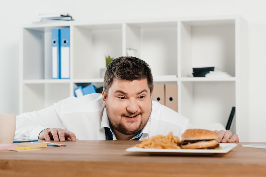 бизнесмен с избыточным весом смотрит на гамбургер и картошку фри в офисе
 - Фото, изображение