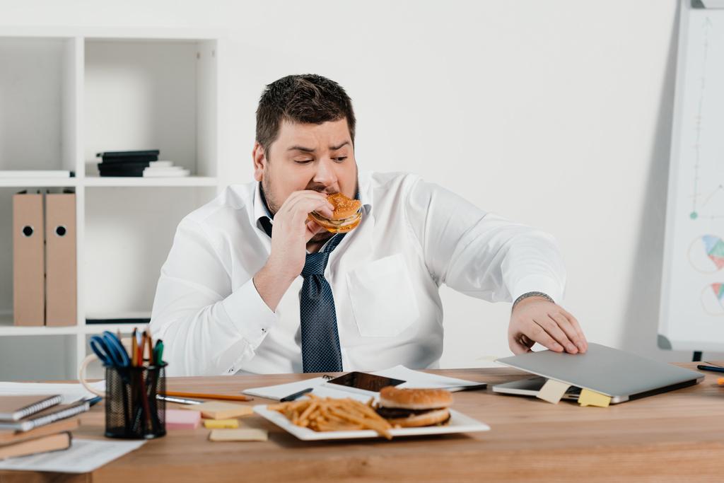 nadwagą biznesmen jedzenie hamburgerów i korzysta z laptopa w biurze - Zdjęcie, obraz
