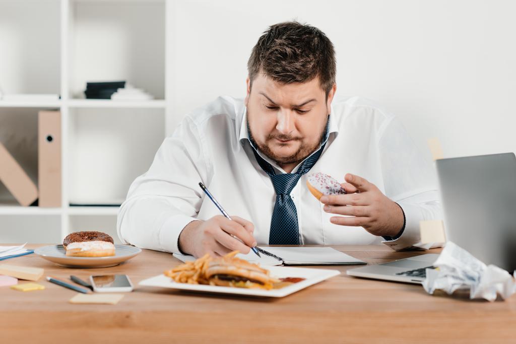 overweight businessman eating donuts, hamburger and french fries while wokring in office - Photo, Image