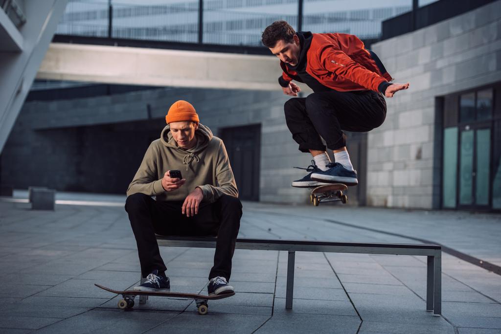 скейтбордист стрибає через лавку, а його партнер сидить на ньому і використовує телефон
 - Фото, зображення