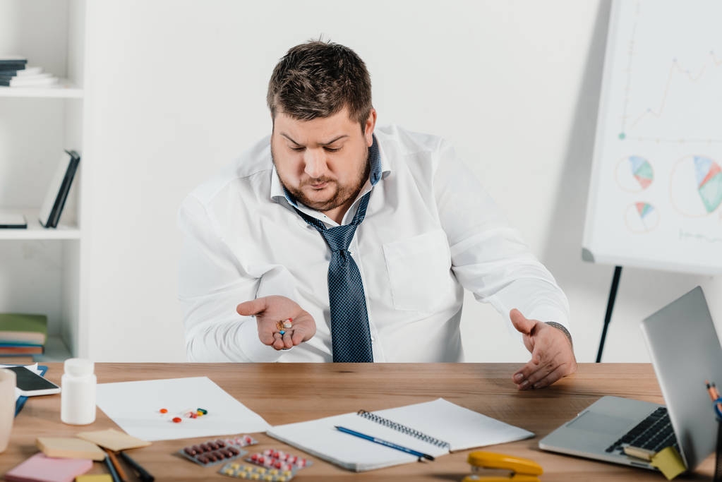 uomo d'affari in sovrappeso seduto sul posto di lavoro e guardando pillole
 - Foto, immagini