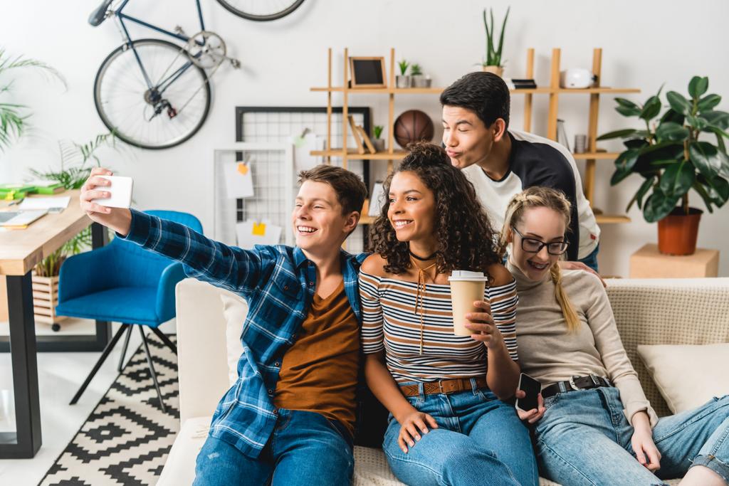 щасливі багатоетнічні підлітки сидять на дивані і приймають селфі зі смартфоном
 - Фото, зображення