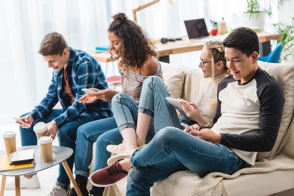 happy multicultural teens sitting on sofa with digital devices  - Photo, Image