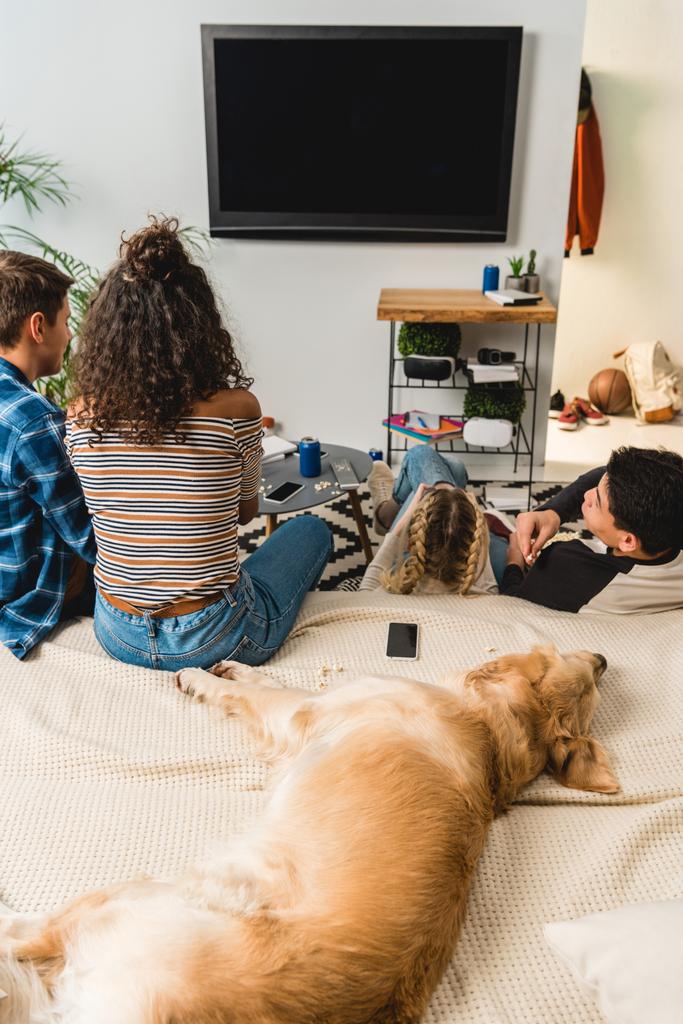 vier tieners tv kijken en hond liggend op bed - Foto, afbeelding