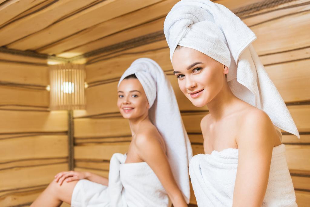 Gelukkig jonge vrouwen bedekt met handdoeken relxing in sauna en camera te kijken - Foto, afbeelding