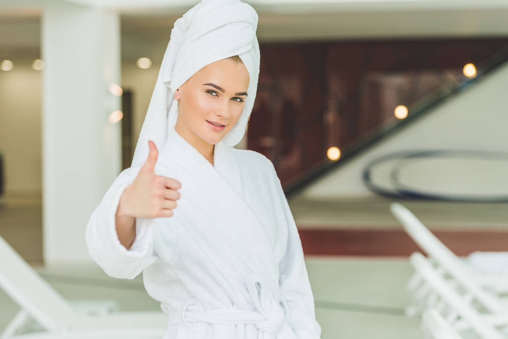 Lächelnde junge Frau im Bademantel im Wellness-Salon zeigt Daumen hoch - Foto, Bild