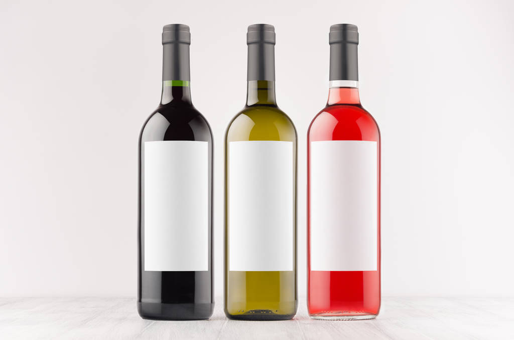 Τρεις κρασιού μπουκάλια - κόκκινο, πράσινο, μαύρο - με κενό οι λευκές ετικέτες σε λευκά ξύλινη σανίδα, ομοίωμα. Πρότυπο για τη διαφήμιση, σχεδιασμός, branding ταυτότητα. - Φωτογραφία, εικόνα