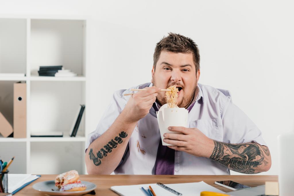 Übergewichtiger tätowierter Geschäftsmann isst Nudeln am Arbeitsplatz im Büro - Foto, Bild