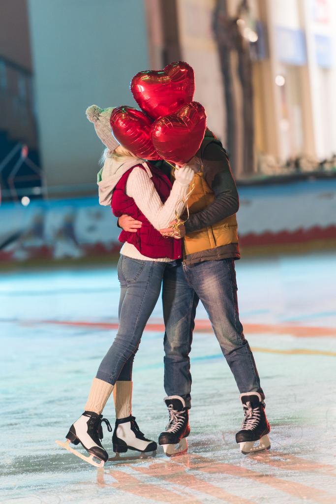 Happy νεαρό ζευγάρι με το αγκάλιασμα στο παγοδρόμιο στο st ημέρα του Αγίου Βαλεντίνου μπαλόνια σε σχήμα καρδιάς - Φωτογραφία, εικόνα