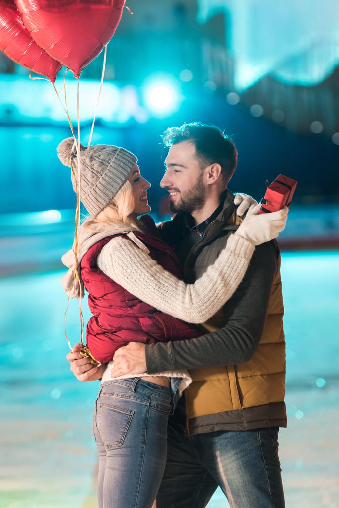 щаслива молода пара з подарунковою коробкою і повітряними кулями у формі серця дивиться один на одного на ковзанах
  - Фото, зображення