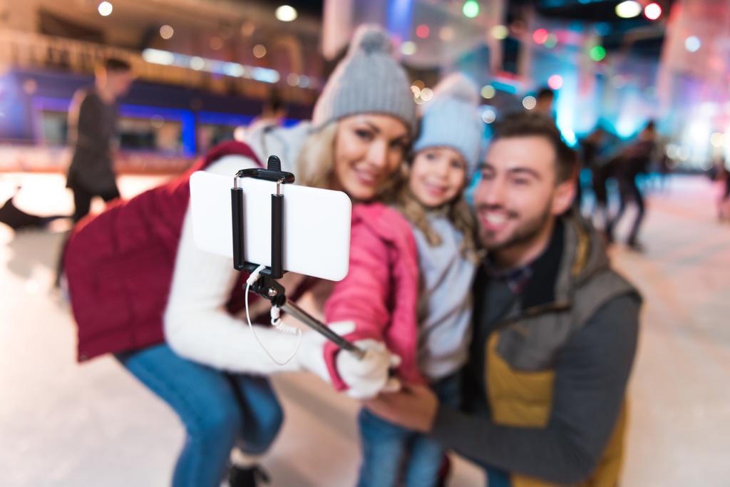 επιλεκτική εστίαση του χαρούμενος νεαρός οικογένειας λαμβάνοντας αυτοπορτρέτα με smartphone στο παγοδρόμιο - Φωτογραφία, εικόνα