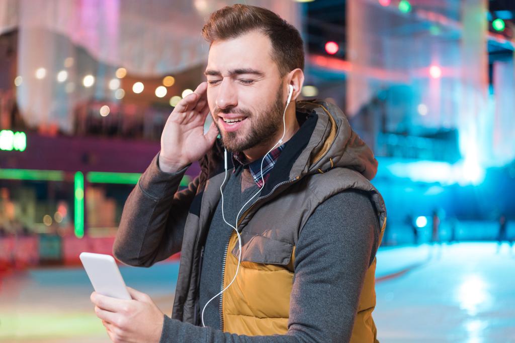 happy young man with closed eyes listening music with smartphone while ice skating on rink - Photo, Image