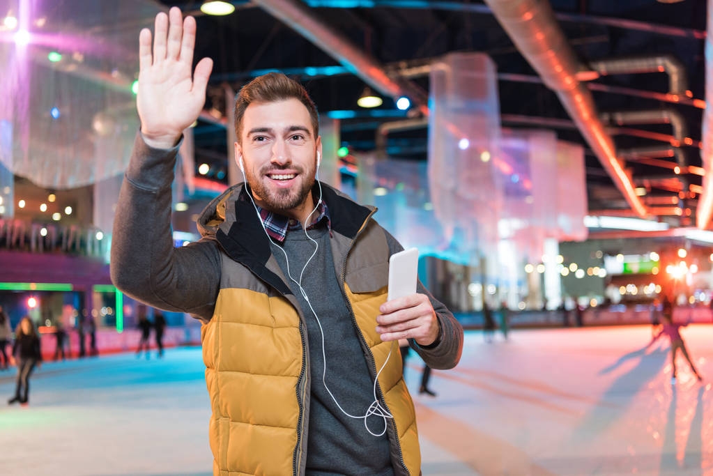 χαμογελαστός νεαρός άνδρας στα ακουστικά κρατώντας smartphone και κουνώντας το χέρι ενώ στέκεται σε παγοδρόμιο - Φωτογραφία, εικόνα