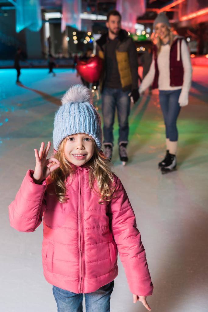 χαριτωμένο μικρό κορίτσι δείχνει εντάξει σημάδι και να χαμογελά στη φωτογραφική μηχανή, ενώ πάγου πατινάζ με γονείς σε παγοδρόμιο - Φωτογραφία, εικόνα
