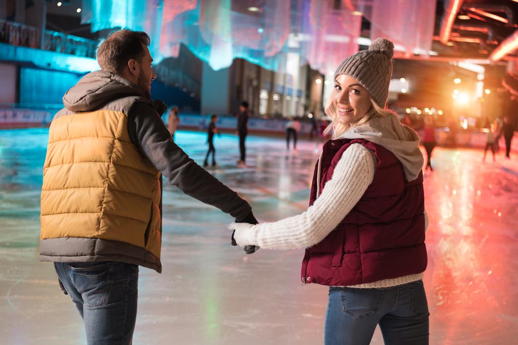 Achteraanzicht van gelukkige jonge paar, houdend handen en schaatsen op de ijsbaan - Foto, afbeelding