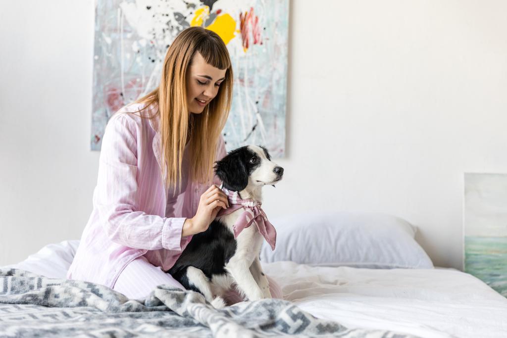 Улыбающаяся женщина в пижаме и милый маленький щенок, отдыхающий на кровати утром дома
 - Фото, изображение