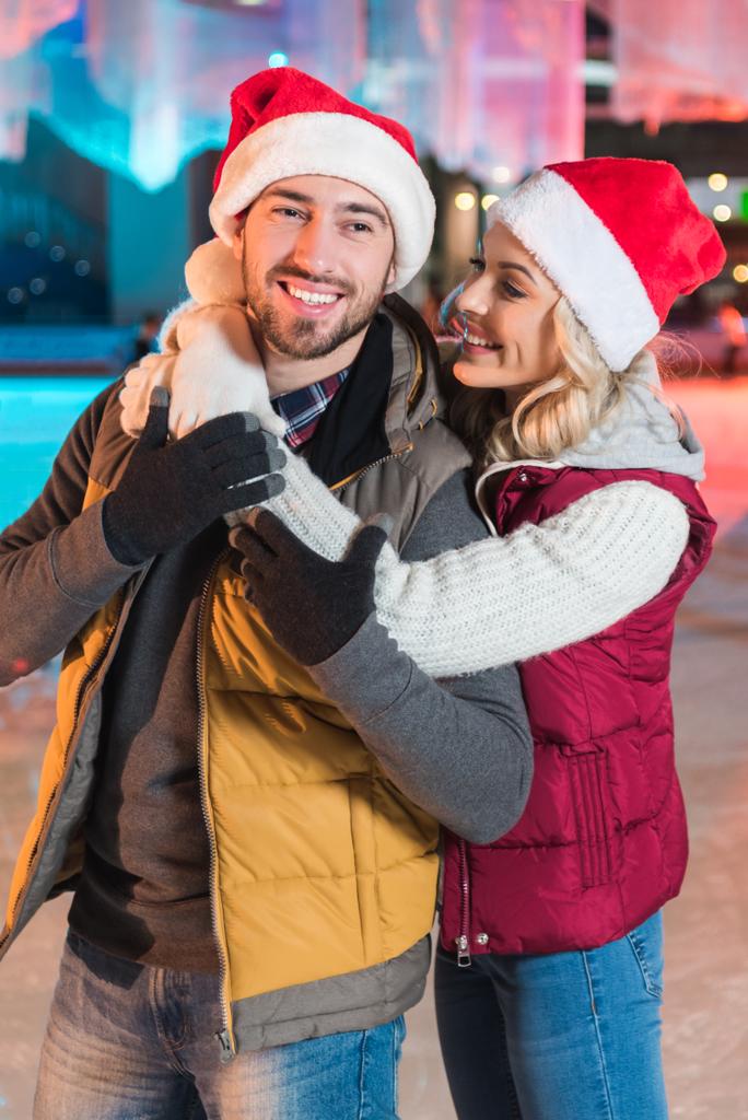beautiful happy young couple in santa hats hugging on rink  - Photo, Image