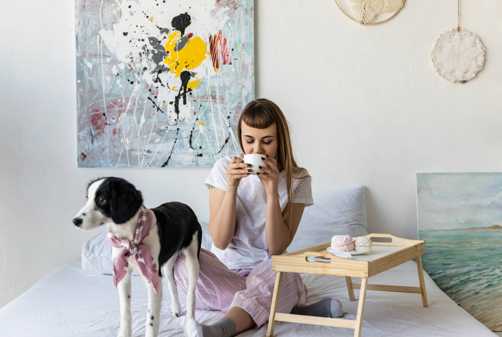 子犬と一緒にベッドで休んでいる間コーヒーを飲む女性 - 写真・画像