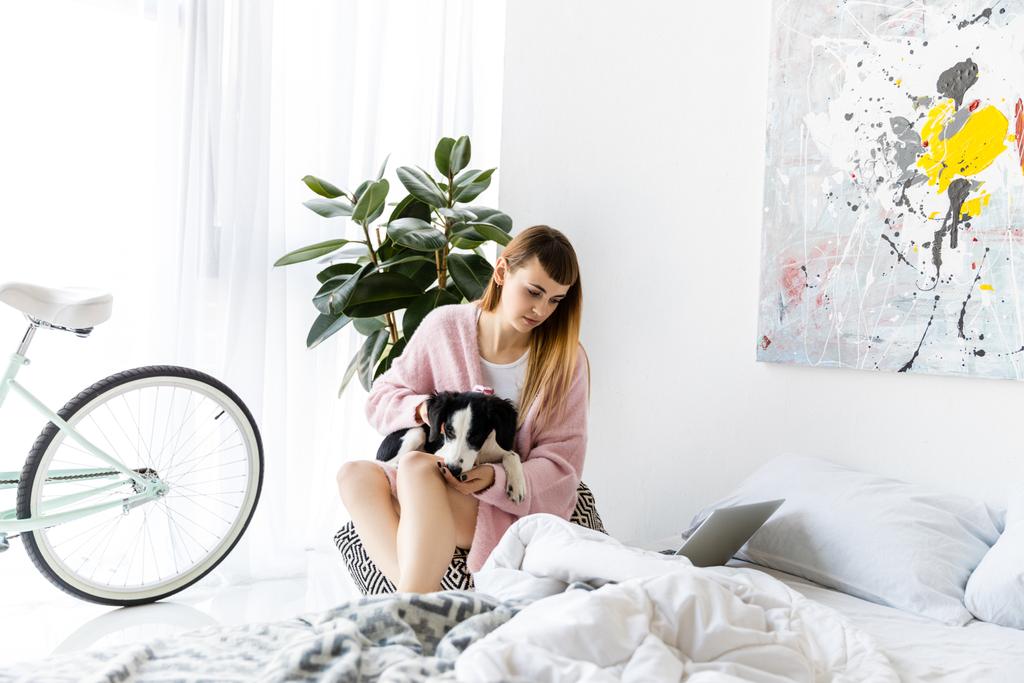 portrait of focused woman with puppy in hands looking at laptop screen at home - Photo, Image