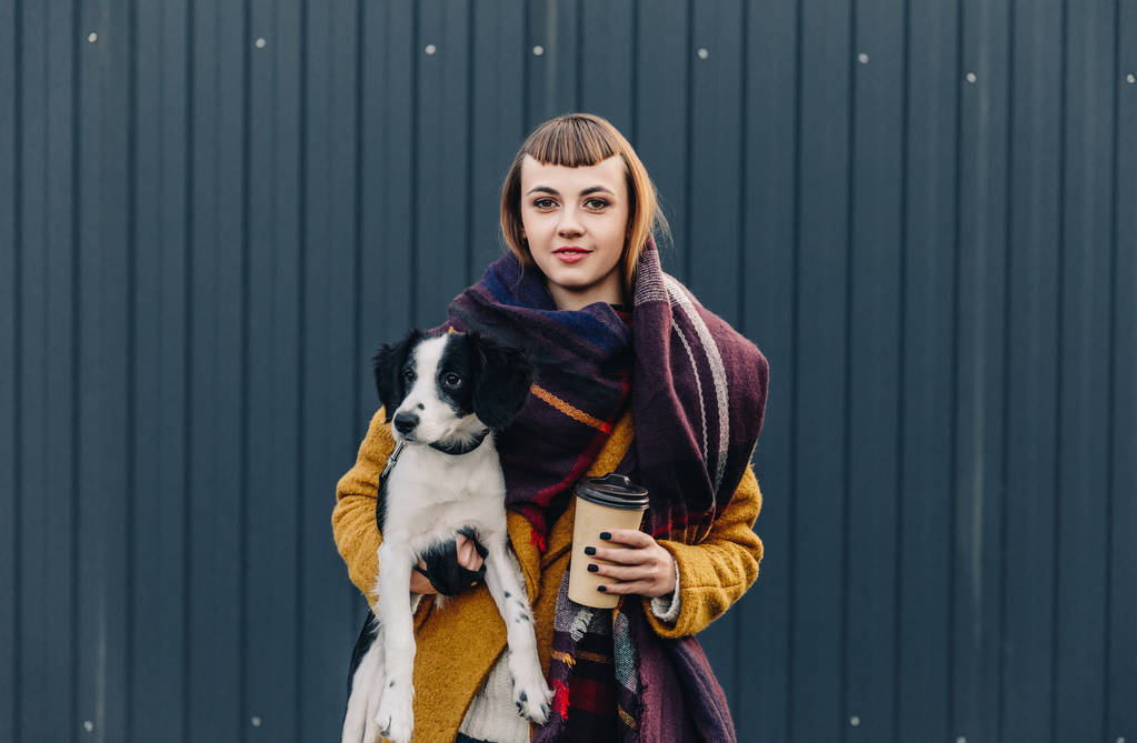 portrét mladé ženy držící štěně a kávu jít do rukou přitom stát na ulici - Fotografie, Obrázek