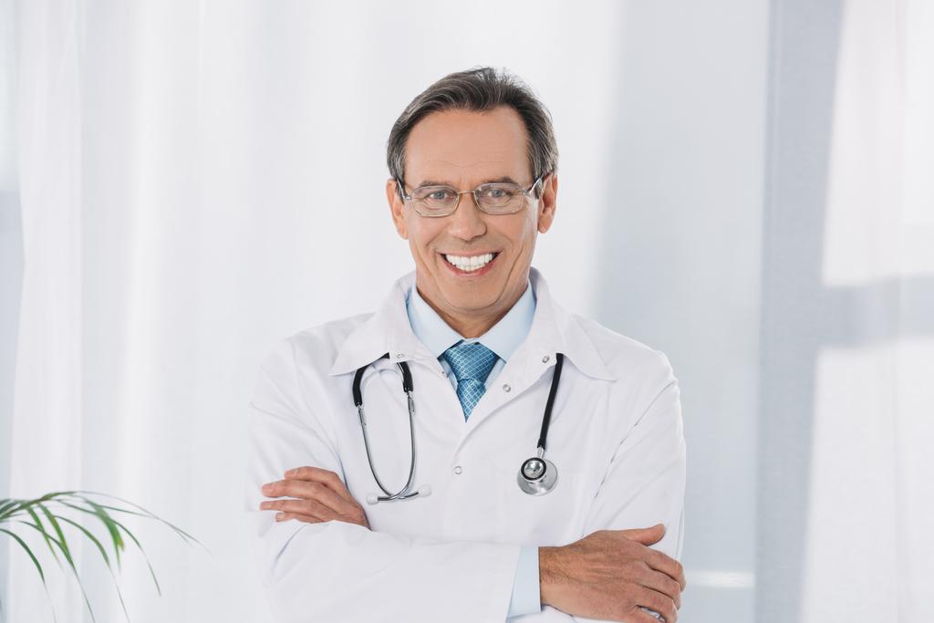 Lächelnder Arzt, der mit verschränkten Händen vor der Kamera steht - Foto, Bild