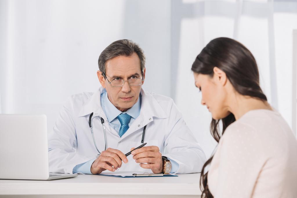 αρσενικό σοβαρό γιατρό και Θηλυκός ασθενής κάθεται στο μιλώντας στην κλινική - Φωτογραφία, εικόνα