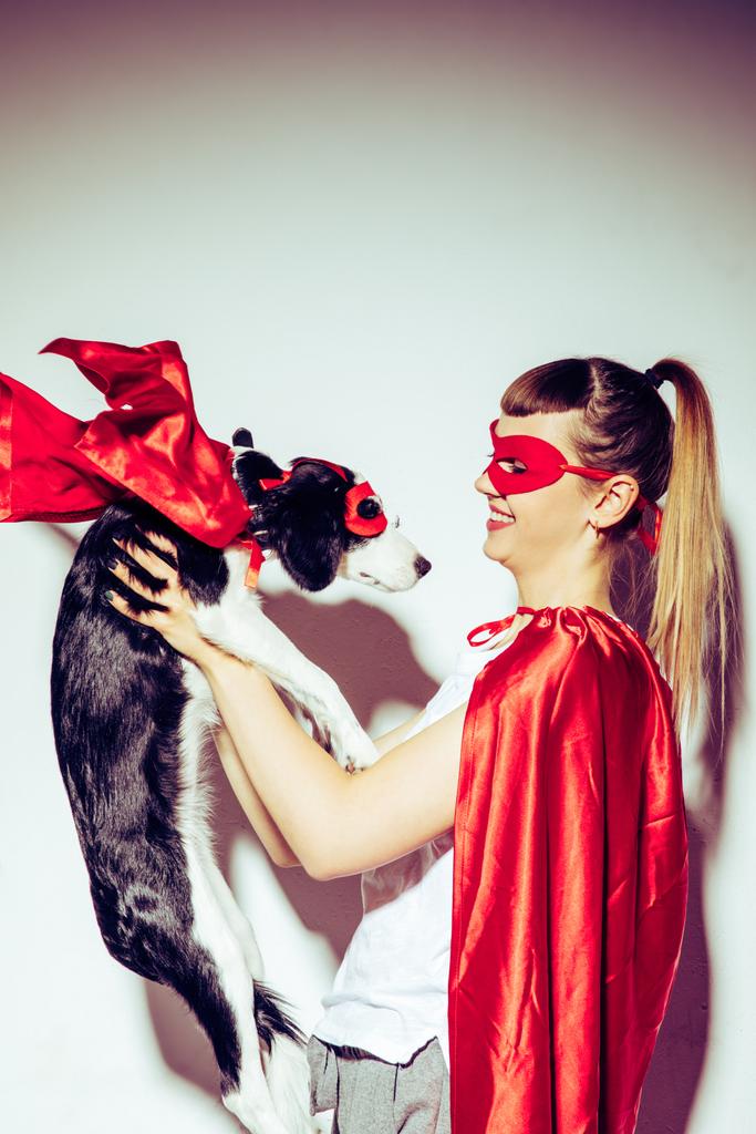 side view of cheerful woman looking at little puppy in superhero costume in hands - Photo, Image