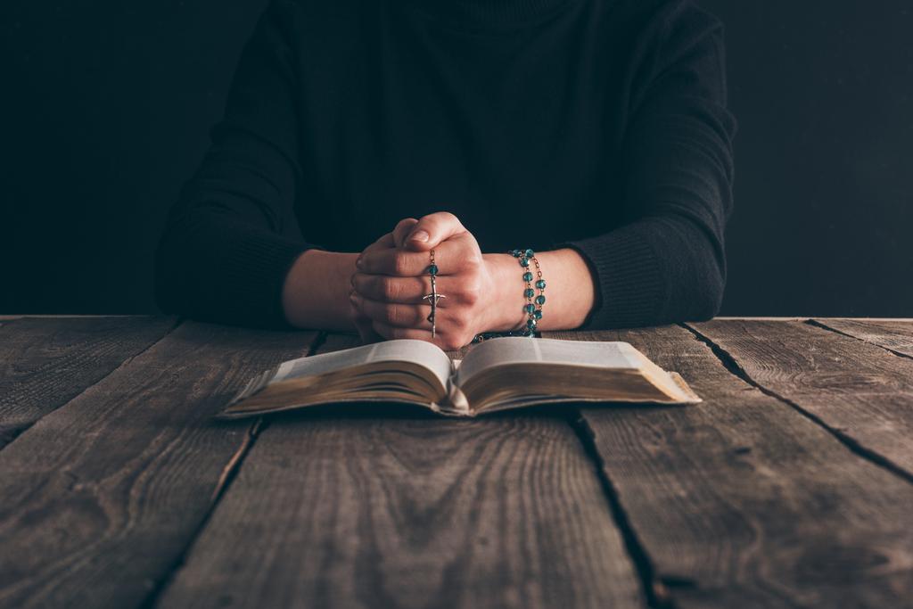 περικομμένη εικόνα γυναίκα που κάθεται στο τραπέζι με το ροδάριο και Αγία Γραφή και προσεύχεται   - Φωτογραφία, εικόνα