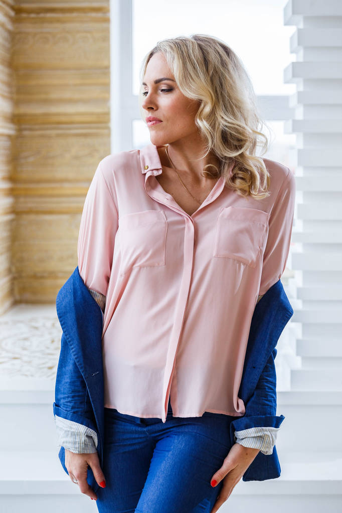 Retrato de beleza jovem loira com cabelo encaracolado, vestindo azul terno casual e camisa rosa em pé pela janela em casa interior
 - Foto, Imagem