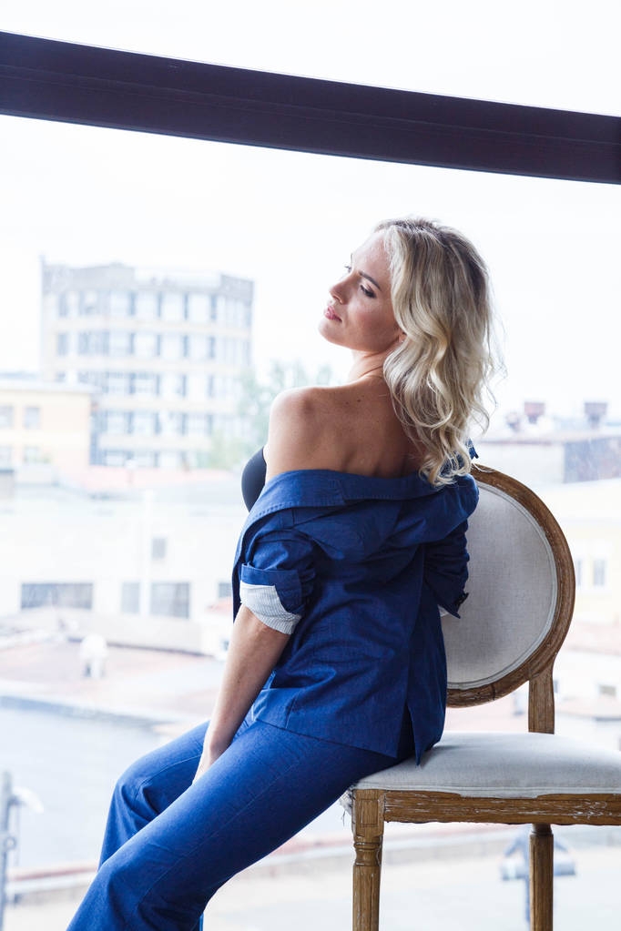 Studioporträt der schönen blonden Frau mit lockigem Haar und stylischem Make-up, trägt blauen Freizeitanzug und blaue Schuhe auf hohen Hügeln, sitzt auf weißem Vintage-Stuhl, verschwommener Blick auf die Stadt im Fenster im Hintergrund  - Foto, Bild