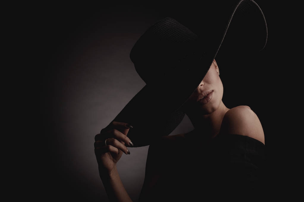黒の広い帽子と黒のドレスでエレガントな女性の劇的な暗いスタジオ肖像画。隠された目. - 写真・画像